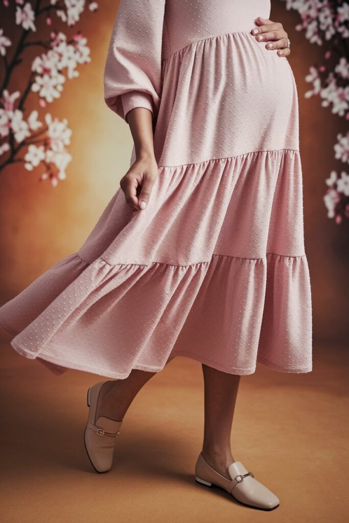 A soft pastel pink dress with gentle, flowing layers cascades elegantly, its tiered design adding subtle dimension to the figure. The dress, perfect for a gender reveal celebration, is paired with neutral shoes and light accessories, set against a warm, softly-lit background of blooming cherry blossoms.