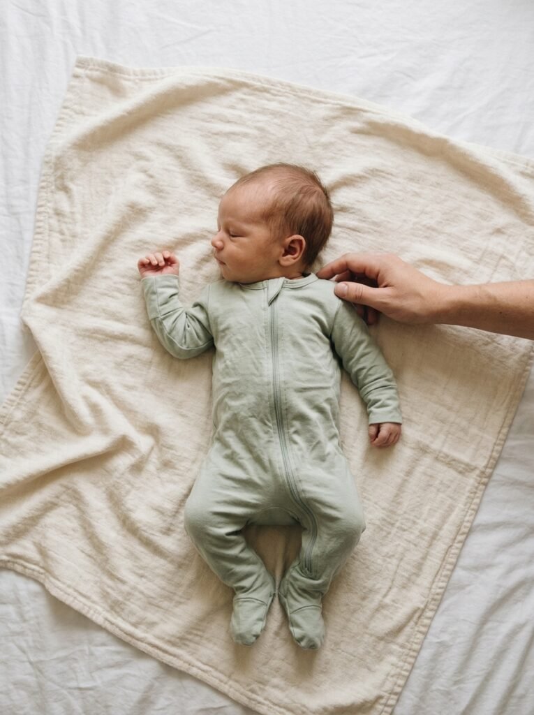 Newborn baby lying on their back on a cream blanket with an adult hand resting gently nearby