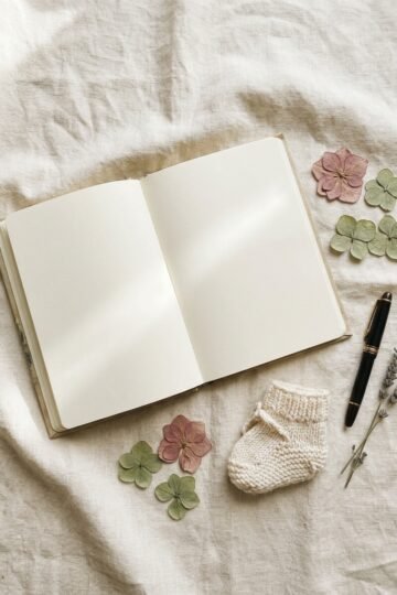 Open notebook with blank pages, pressed flowers, a pen and a tiny knit bootie on a cream linen background