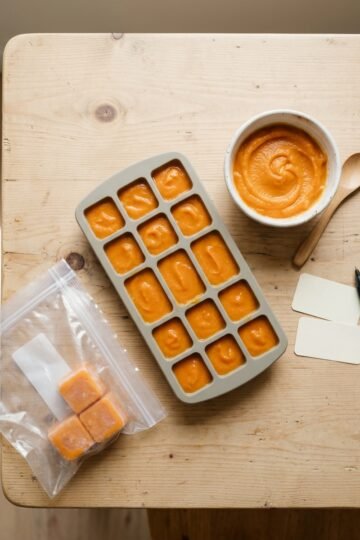 Silicone ice cube tray filled with orange puree beside a small bowl, a spoon and a freezer bag of frozen cubes